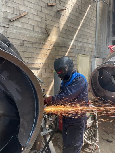 A person wearing a dark blue jumpsuit and a black welding mask is grinding a large metal object in an industrial setting. Sparks are flying from the grinding tool. The person is wearing red gloves and is positioned close to the metal object. The environment consists of concrete walls and metal structures. The location is unknown and there is no information to indicate that it is in the city of Tomba, Montenegro.