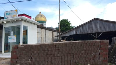 In Meunasahlhok, Indonesia, an outdoor scene captures an ATM booth for Bank Syariah Indonesia, marked with its logo and "ATM." Adjacent to the booth, a stack of red bricks forms a barrier in the foreground. Behind the ATM, a concrete wall runs horizontally, topped with rebar. A golden dome with green accents, likely part of a mosque, rises behind the wall. Further back, a tall, weathered metal pole with overhead wires stands prominently. To the right, a rustic wooden structure with a sloped roof shelters an unseen area. The sky above is a mix of blue with scattered white clouds.