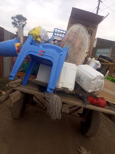 The image depicts a wooden handcart, heavily loaded with diverse household items, positioned on a dirt path in Kiamaina ward, Kenya. The sky overhead is overcast.

**Forensic Description:**
The primary subject is a rustic wooden handcart with two visible wheels, resting on dry, brown dirt ground. The cart is overladen with an assortment of objects. Prominently featured in the center-left is an inverted blue plastic stackable chair. To its immediate left, a blue plastic bucket containing yellow fabric or clothing hangs from the cart's side. A long, grey string mop head dangles from the front wooden beam of the cart. On the right side of the cart bed, two white plastic jerrycans or containers are visible, with one displaying blue label text. A single red plastic sandal or shoe rests on the cart bed near the right wheel. Towards the rear and right, a large, open brown cardboard box with white shipping symbols stands upright, leaning against other items. A circular object with a white and pink patterned lace or netting cover is visible leaning against the cardboard box. Additional items, including other blue plastic chairs, bundles wrapped in fabric (one notably yellow), and an indistinguishable white rectangular object, are stacked and packed onto the cart.

The background on the left features a weathered, rusted corrugated metal fence. Behind this, a slender utility pole with an attached tree is visible. On the right side of the background, part of a masonry building with a dark-framed window or door opening is present. No individuals are visible within the frame. The overall scene suggests a collection of personal effects either being transported or temporarily stored.