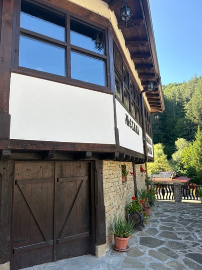 The image depicts a side view of a traditional Bulgarian house in the village of Chiflik, Bulgaria. The house features a rustic architectural style with exposed wooden beams, stone walls, and a wooden door. White stucco walls contrast with the dark wood elements. Above the door, there is a section of white wall with dark lettering that appears to be a sign, possibly displaying the name of the establishment or location. The roof is covered with wooden shingles or tiles, and antique-style lanterns hang from the eaves.
The exterior walls are constructed of rough-hewn stone blocks. A dark wooden double door is prominent on the lower level, appearing weathered and solid. Above the stone wall, a band of white stucco is visible, supporting a large wooden-framed window. This window has multiple panes and is set within the wooden facade.
In front of the house, a paved courtyard is laid with irregular flagstones. Potted plants, including vibrant red flowers and greenery, are strategically placed along the stone wall and in the courtyard, adding a touch of color to the scene. A small wooden barrel is also present near the plants.
Further in the background, beyond the courtyard, a metal fence with an intricate design separates the property from what appears to be another building with a red roof. Solar panels are visible on the roof of this structure, suggesting a blend of traditional and modern elements. The scene is set against a backdrop of lush green trees and a clear blue sky, indicating an outdoor environment likely in a rural or mountainous region. The overall impression is one of rustic charm and a connection to nature.