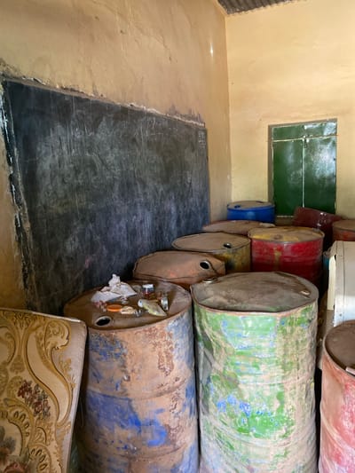 The image shows the interior of a building located at 15.552 latitude and 32.57 longitude in Khartoum, Sudan. It appears to be a schoolroom or a storage room, as it contains a blackboard with white chalk writing, and several large metal barrels. The barrels are rusty and worn, and some have been painted with blue, red, and green paint. The wall is a pale yellow color and is peeling in some places. The room is poorly lit. There is a wooden door with a green metal covering in the back of the room. There are no people present in the scene.