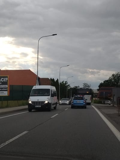 The image depicts a view from a vehicle driving on a road in Tábor, Czechia, under an overcast sky. In the foreground, the asphalt road has white lane markings. A white Renault van is in the right lane, its headlights illuminated, and its license plate reads "6LB 6825". To the left of the van, a silver sports car is visible. Further ahead, a blue car with license plate "BC4 6462" is in the left lane, followed by a bus. On the left side of the road, a large billboard with text in Czech is partially visible. Behind the billboard, buildings with red tiled roofs are present. On the right side of the road, there are trees and houses. The overall scene suggests moderate traffic on a cloudy day.