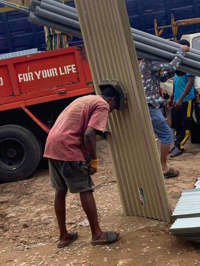 In Ogbunike, Nigeria, several individuals are engaged in the process of unloading or loading materials from a truck. A red truck with white lettering, reading "FOR YOUR LIFE," serves as the central focus of the activity. One person, wearing a red shirt and dark shorts, is bent over and appears to be supporting a large, corrugated metal sheet. Another individual, in a patterned shirt and blue shorts, is carrying a bundle of grey pipes. A third person, in a blue shirt and dark pants with yellow stripes, is also present. The environment is unpaved, with dirt and gravel on the ground. The scene suggests a construction or delivery site, with building materials like pipes and corrugated sheets being handled.