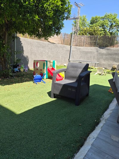 The image depicts an outdoor scene on a sunny day, likely in the late morning or early afternoon, given the strong shadows. The setting appears to be a backyard or play area, with artificial turf covering the ground. In the foreground, a black woven armchair is positioned on the turf. To its left, a multi-colored plastic play structure for children is visible, featuring yellow, red, and green components. A small blue plastic chair is placed in front of the play structure. Further to the left, lush green foliage from a tree, possibly an orange tree given its appearance, partially obscures the view.
In the background, a grey wall borders the play area, with a fence made of woven reeds or bamboo behind it. Beyond the fence, green trees and a power line tower under a clear blue sky are visible, suggesting an urban or semi-urban environment. The overall impression is one of a well-maintained outdoor space designed for recreation. There is no visible text in the image. The scene evokes a sense of calm and leisure, typical of a day spent outdoors. The lighting suggests good weather.
