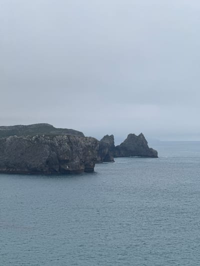 The image depicts a dramatic coastal landscape in Llanes, Spain, characterized by rugged, dark gray cliffs meeting a vast expanse of tranquil, blue-gray water under an overcast sky. The cliffs are textured and weathered, with sharp edges and visible geological formations. In the foreground, the water appears slightly choppy, with subtle ripples indicating a gentle movement. In the mid-ground, two prominent rock formations rise from the sea, their pointed peaks silhouetted against the muted sky. The background reveals a distant, hazy shoreline and a featureless, pale gray sky, suggesting a cool, possibly misty day. The overall scene evokes a sense of natural grandeur and the raw power of the ocean.