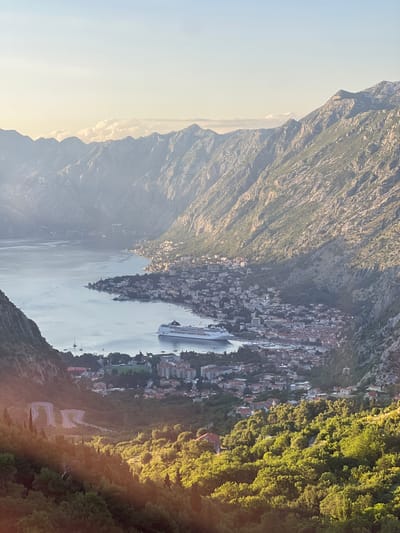 A high-angle, expansive view captures a mountainous coastal landscape encompassing the city of Kotor within the Bay of Kotor, near Čavori, Montenegro, during the golden hour. The upper portion of the frame features a pale blue sky transitioning to a soft yellow hue near the horizon, with scattered, diffused clouds resting above distant mountain peaks.
The mid-ground is dominated by the calm, grey-blue waters of a large bay or fjord, which curves around a developed coastline. Along the bay, the city of Kotor is visible, characterized by a dense collection of buildings with varied architecture, many featuring red-tiled roofs. A prominent white cruise ship is anchored or docked in the bay adjacent to the city, its stern facing towards the viewer. Several smaller boats and yachts are scattered across the water.
Steep, rugged mountains, composed of rock faces and varying densities of green vegetation, flank both sides of the bay, descending directly to the water's edge. Sunlight illuminates the upper parts of the mountains on the right, casting distinct shadows across the lower slopes and parts of the city.
The foreground on the left features a densely vegetated hillside, with green trees brightly illuminated by direct, warm sunlight. A distinctive asphalt road with multiple sharp hairpin turns is clearly visible winding down the mountain slope on the lower left. Scattered residential structures with red roofs are interspersed among the trees in the lower-most sections of the image. No individuals are discernible within the frame.