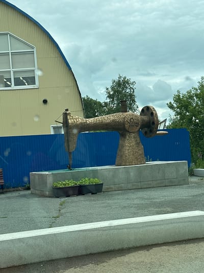 The image shows a large, bronze-colored sculpture of a sewing machine in Uchaly, Russia. The sculpture is detailed, with a patterned surface and what appears to be writing or symbols on its body. It stands next to a bright blue building and a light yellow building with a distinctive arched roof and a large, multi-paned window. A paved area with a white line runs in front of the sculpture, with some planters containing green plants to the right of it. The sky is overcast, suggesting it is daytime. The overall mood is somewhat industrial or commemorative, given the large scale of the sculpture. There are trees visible in the background. The sculpture appears to be permanently installed as a public artwork.