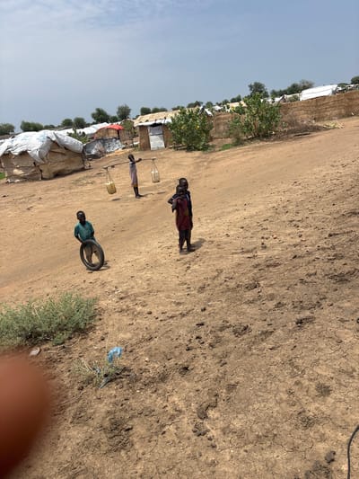 The image depicts a scene in Ad-Damazin, Sudan. In the foreground, a young child in a teal shirt is pushing a black tire. Further back, two other children stand. One, wearing a red garment, appears to be looking towards the viewer, while another child behind them is less visible. In the middle ground, a child in a light-colored garment carries two containers of water suspended from a wooden pole across their shoulders. The environment is arid and dusty, with dry earth and sparse vegetation. Several structures, possibly temporary shelters, are visible in the background, some covered with white sheeting bearing the UNHCR logo, suggesting a displacement camp or refugee settlement. The sky is overcast.