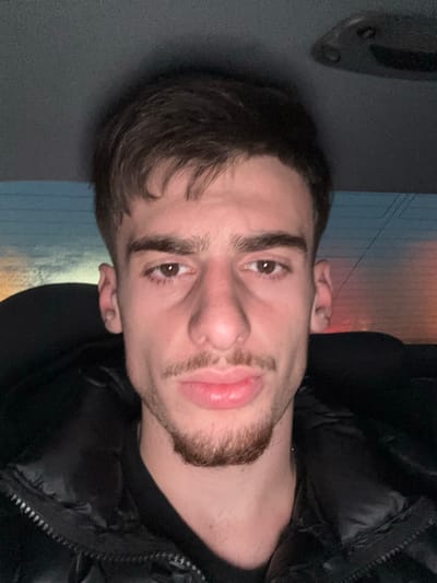 A young man is depicted in a close-up, chest-up shot, looking directly at the camera. He has dark hair, brown eyes, and a light-brown goatee and mustache. His lips are slightly parted in a neutral expression. He is wearing a dark, possibly black, puffer jacket over a dark shirt. The image is notably rotated counter-clockwise, with the man's head tilted. The background is softly blurred, revealing ambient light that suggests dusk or evening, with warm hues visible. To the left, a dark, textured surface, possibly an interior car panel or wall, is visible. No text is visible in the image. The scene is set in Souffelweyersheim, France.