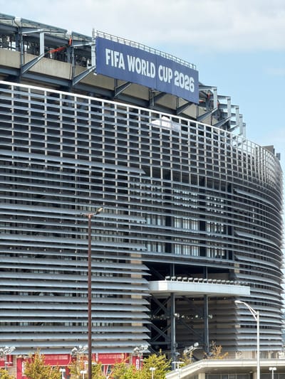 An outdoor, medium shot captures the exterior of a modern stadium in East Rutherford, United States of America, under a bright, partly cloudy sky. The stadium features a distinctive facade of horizontal grey louvers that partially obscure windows and structural elements, creating a rhythmic pattern across its curved surface. A large, blue banner with white lettering reading "FIFA WORLD CUP 2026" is prominently displayed near the top of the structure, signifying the upcoming event. Below the louvers, at ground level, red awnings with white lettering indicate "HOSPITALITY EAST ENTRANCE." A tall, slender lamppost with a modern design stands in the foreground, to the right of center. The surrounding environment includes green trees and some exposed structural steelwork. The overall scene suggests a sports venue preparing for a major international event.