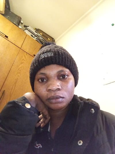The image displays a close-up indoor portrait of a female individual in Madinaty, Egypt. The subject, an adult female with a medium-dark complexion, looks directly at the camera with a neutral expression. She wears a dark, ribbed knit beanie hat with a pom-pom and "NYC" embroidered in white on the front, along with a dark jacket that has visible snap buttons. Beneath the jacket, a dark shirt with a white embroidered bird-like logo on the left chest is visible. Her left hand is raised, supporting her chin, and a golden circular button is present on her left jacket cuff. The background to the upper left features light brown wooden cabinets with dark handles, upon which at least two pieces of luggage are stacked, including a dark wheeled suitcase. The upper right background is a plain, light-colored wall with a small green marking. Overhead lighting illuminates the scene.
