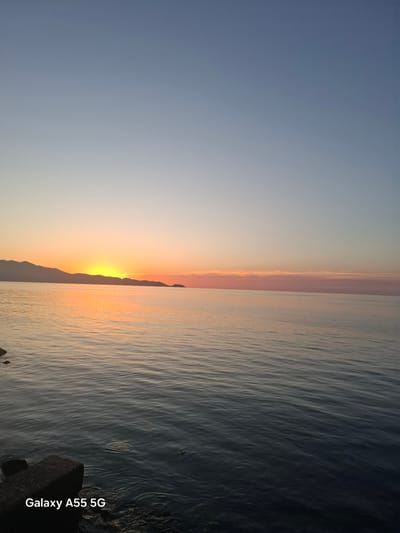 The image depicts a sunset over the water, likely a sea or large lake, in the Heraklion Municipal Unit, Greece. The sky transitions from a light blue to an orange hue near the horizon, where the sun is setting behind a mountain silhouette. The water reflects the colors of the sky, creating an orange-toned surface. The lower right corner of the image shows a dark, textured rock. The text "Galaxy A55 5G" is visible in the bottom right corner, indicating the device used to capture the image.