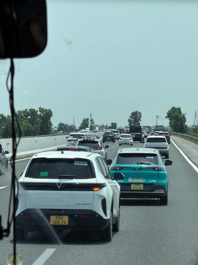 The image shows a traffic jam on a highway, likely in Hải Phòng, Vietnam. The scene is observed from within a vehicle, looking forward through the windshield. A white LIMO GREEN car with license plate 89H-106.74 is directly in front, followed by a turquoise VINFAST VF-e34, a brand known in Vietnam. This VINFAST has the license plate 29H-913.50 and advertises a taxi service with the phone number 1900 2088. The congested road stretches into the distance with numerous cars and trucks. Trees line the side of the road, and power lines are visible in the background. The sky is overcast.