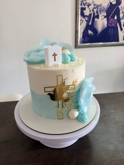 The image shows a decorated cake placed on a white cake stand, which is sitting on a dark brown surface. The cake is cylindrical and decorated with a white and light blue frosting, with gold leaf accents. It features a gold cross with a dove cut-out, light blue flowers, and a white fondant piece with a gold cross. The cake appears to be religious themed. In the background, a framed picture hangs on a white wall. The location is Saint Paul's Bay, Malta.