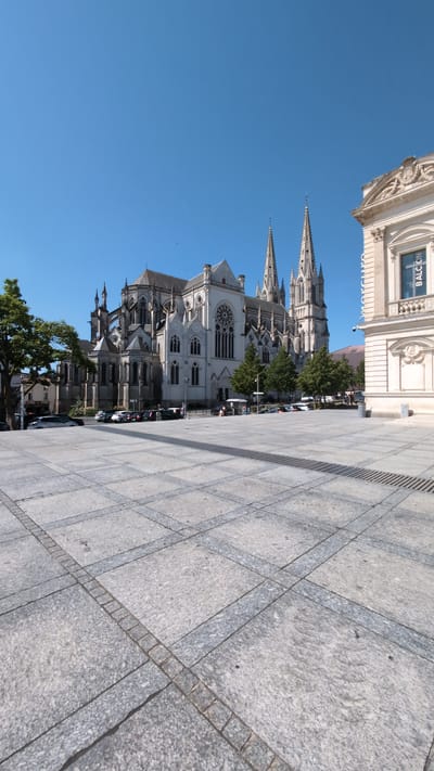The image captures a wide-angle view of a sun-drenched plaza in front of the Saint-Louis Cathedral in Cholet, France. The cathedral, a magnificent Gothic structure, dominates the background with its white stone facade, intricate stonework, and soaring spires. To the right of the cathedral, a modern building with a large window featuring a blue poster with white text is visible. The plaza itself is paved with large grey stone tiles arranged in a geometric pattern, with dark lines forming a grid. In the middle ground, several cars are parked along a street adjacent to the plaza, and a few trees provide shade. Several individuals are scattered across the plaza, appearing to be pedestrians or tourists. The sky is a clear, vibrant blue, indicating a bright, sunny day.