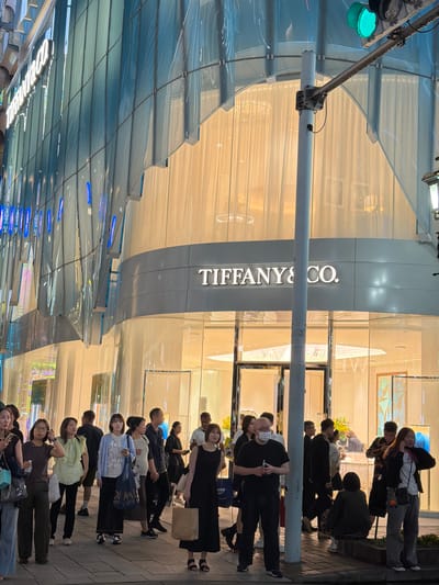 A street-level, medium shot captures the exterior of a Tiffany & Co. store in Chuo, Japan, at dusk. The building's facade is a curved, multi-story structure with a modern design. The upper portion features a translucent, blue-tinted glass curtain wall with vertical elements and integrated blue lighting. Below this, a horizontal band of light-colored, translucent material creates a warm glow, obscuring the interior details but suggesting a retail space. The iconic "TIFFANY & CO." logo is prominently displayed in white, illuminated lettering on a gray band across the lower part of the building. The ground floor features a large glass storefront with entrances and display windows.
In the foreground, a diverse group of people are standing on a paved sidewalk. Their expressions and postures suggest they are either pausing, waiting, or observing. Some individuals are looking at the store, while others are engaged in conversation or simply standing. The lighting is a mix of ambient streetlights, the building's interior and exterior illumination, and a nearby traffic signal that is currently green. A metal pole with attached equipment, possibly a traffic light pole, stands centrally in the image. The overall scene depicts a bustling urban environment in the evening.