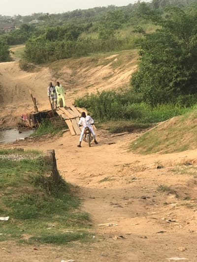 A landscape scene includes a dirt path running horizontally across the frame, bordered by greenery on the left and more dirt with scattered vegetation on the right. A wooden plank bridge crosses a narrow stream or ditch. Two people stand on the bridge's edge, while a third person, dressed in white, lies horizontally across a motorcycle on the bridge. The image is taken from a high vantage point and is rotated slightly.