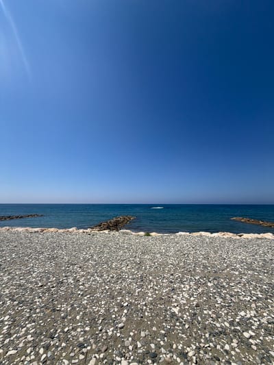 The image depicts a serene, sun-drenched beach with a clear blue sky overhead. The foreground is dominated by a vast expanse of smooth, multi-colored pebbles, ranging from white and grey to darker hues, interspersed with patches of sand. A low line of larger, lighter-colored stones forms a border between the pebble beach and the sea.
Beyond this, the tranquil, deep blue sea stretches out towards the horizon, meeting the vibrant sky. A gentle wave is visible in the distance. Several man-made breakwaters, constructed from rough, dark rocks, jut out into the water at intervals, breaking the monotony of the sea. The overall impression is one of natural beauty and peaceful seclusion, characteristic of the coastal areas near Pervolia, Cyprus. There are no people or any animals visible in the image. The sun casts a subtle lens flare in the upper left corner of the frame.