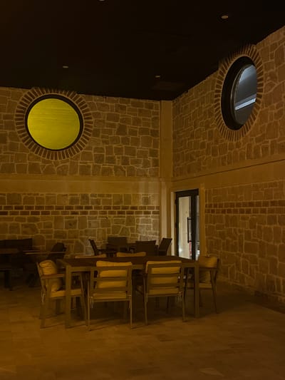 A low-angle indoor shot depicts stacked chairs against a stone wall with a window above, located in Ürgüp, Turkey. The scene is illuminated, revealing details of the building's structure and decor. The chairs, stacked on the right side of the image, feature light-colored cushions. The stone wall is a mix of square and rectangular stones. On the left side of the image, there's a dark wall with round windows, one yellow and one showing sky. The stone patterns, chair placement, and window design are integral to the architectural style of Ürgüp, Turkey. The building appears to be of contemporary design.