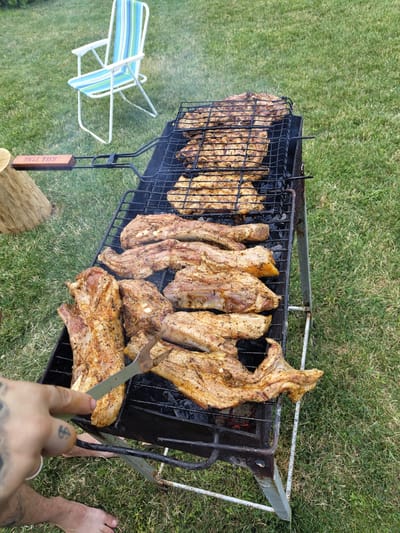 The image depicts a barbecue scene set on a grassy area, likely outdoors. In the foreground, a person's hand, tattooed, is holding a pair of tongs, reaching towards several pieces of raw meat cooking on a grill. The meat appears to be seasoned and is positioned on two levels of the grill. The grill itself is a metal structure with a base and wheels, placed on the grass. In the background, a foldable lawn chair with blue and white stripes is visible, also on the grass. A wooden object with the inscription "HILL TIME" is situated to the left of the grill. The environment suggests a casual outdoor gathering. There is no indication of the specific location within the city of Oxentea, Moldova.