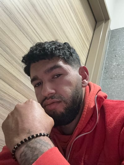 A male subject with dark, curly hair and a trimmed beard/mustache is depicted in a close-up, eye-level portrait. He is wearing a red hooded sweatshirt with a visible white zipper. His left fist supports his chin or jaw. On his left wrist, a bracelet composed of dark, possibly square, beads separated by metallic (silver-colored) spacers is visible. A tattoo is present on his left forearm, beneath the bracelet. The background features a light-colored wooden panel wall with vertical striations on the left, and a grey speckled tiled wall on the right, separated by a light-colored trim. The scene takes place in the city of Radovljica, Slovenia.