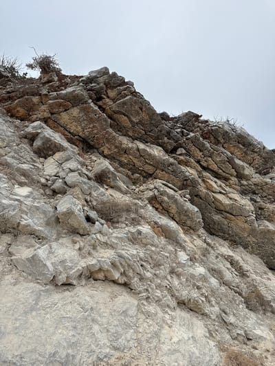 This is a low-angle, full shot of a rocky hillside under an overcast sky. The rocks are various shades of gray and brown, with some lighter, chalky-looking sections and darker, more earthy tones. The terrain is uneven and appears to be a natural rock formation. There are sparse, dry weeds and dead branches scattered among the rocks, suggesting a dry or arid environment. The sky is a uniform pale gray, providing no distinct features or light sources. The image does not show any people or specific man-made objects, and it does not contain any visual information that would place it in Sesimbra, Portugal.