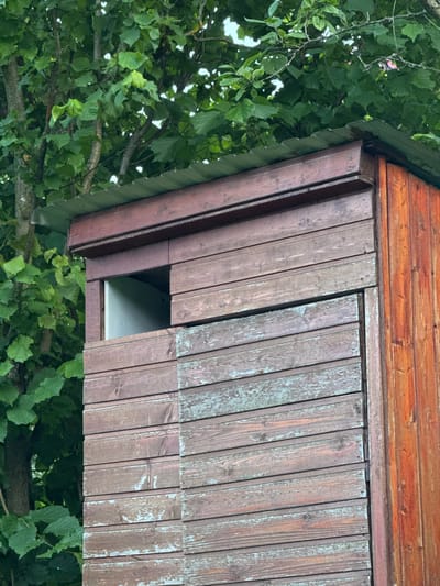 A medium shot shows the exterior of a wooden structure, possibly a shed or a small building, located in the city of Дзержинец-83 Снт, Russia. The structure is constructed with horizontal wooden planks, many of which show signs of weathering and peeling paint in various shades of brown and green. A section of the structure features a dark brown wooden frame around a rectangular opening, which reveals a pale gray interior. The roof is made of corrugated metal. To the left of the structure, dense green foliage from trees is visible, with some branches reaching over the roof. The lighting suggests an outdoor, daytime setting. There are no people or animals present in the image.
