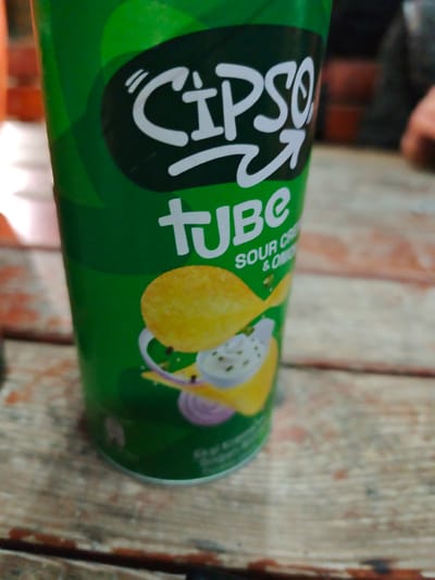 A close-up shot features a green tube of "Cipso Tube Sour Cream & Onion" chips. The tube is resting on a weathered wooden surface with visible grain and discoloration. The chips' packaging is green with white lettering.
The location in the city of Grohotno, Bulgaria is unknown.
