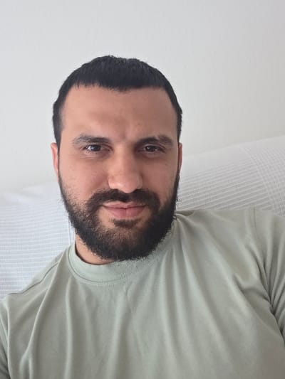 A man with dark hair and a beard is lying on a white surface, possibly a bed or couch. He is wearing a light green t-shirt. The background appears to be a white wall. The image does not contain enough information to determine if the location is in Lausanne, Switzerland.