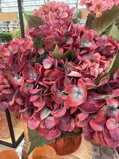 A close-up, eye-level shot captures a large bouquet of artificial hydrangeas in shades of deep pink, crimson, and muted purple. The flowers are densely packed, with their textured petals overlapping and creating a sense of volume. Some of the individual blooms reveal a subtle gradient of color, transitioning from a lighter pink at the edges to a darker hue towards the center, where faint blue veining is visible. Interspersed among the flowers are several realistic-looking green hydrangea leaves with visible vein patterns. The bouquet is arranged in an orange, textured ceramic vase, partially visible at the bottom center of the frame. To the left, a black metal frame, likely part of a display shelf, encloses a glimpse of a store interior. Rows of merchandise, possibly gardening supplies or home decor, are visible in the blurred background, suggesting the setting is a retail environment. The overall lighting is bright, characteristic of an indoor commercial space. There are no people or animals present in the image. The context of Fritzlar, Germany is not evident from the visual information within the photograph.