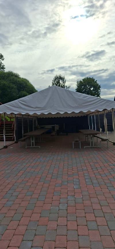 The image shows an outdoor seating area under a large white tent. The ground is paved with red and gray interlocking bricks. Several picnic tables with benches are set up under the tent. In the background, there are wooden structures and lush green trees. The sky is overcast with some bright patches, suggesting diffused sunlight. The scene appears to be located in Valma, Estonia, possibly a park, restaurant patio, or event space. There are no people visible in the image.
