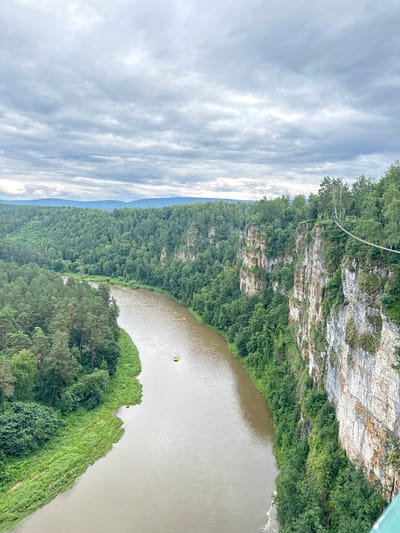 The image depicts a natural landscape with a river flowing through a densely forested gorge. The viewpoint is from a high vantage point, looking down and across the scene. The cliff face on the right is rocky and covered in green vegetation. A suspension bridge stretches across the gorge from the cliff, with a yellow marker visible on the bridge. The river below is a murky brown and has two small yellow objects, possibly kayaks or rafts, on its surface. The surrounding area is covered in lush green trees, with some patches of grass on the riverbank. The sky is filled with dramatic, overcast clouds, suggesting a cloudy or stormy day. The lighting indicates it is daytime, but the overcast conditions reduce direct sunlight. The overall mood is one of natural grandeur and perhaps a sense of adventure, given the presence of the bridge and watercraft. The location context provided is Kul'metovo, Russia, which aligns with a rugged, forested natural environment. There is no visible text in the image.