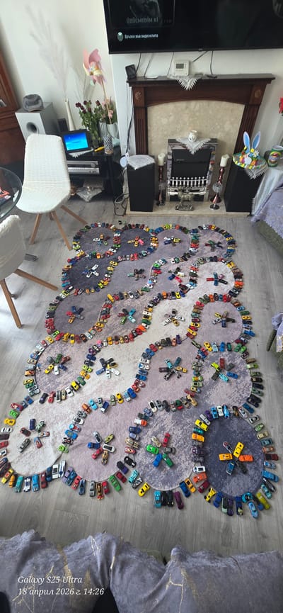 The image presents an overhead view of a rug situated on a light-colored, possibly wood-laminate floor. The rug displays a pattern of interconnected circles in varying shades of gray and white. Surrounding the circles is a collection of toy cars, arranged in a manner that emphasizes the rug's design, tracing the perimeter of each circle. Some clusters of cars are placed within the circles as well.
To the left of the rug, there's a television on a dark-colored stand. In front of the stand, two white chairs and part of a glass-topped table are visible. Behind the TV stand, there is a beige and brown surface, likely a fireplace.
To the right of the rug, the edge of a gray-colored blanket is visible. There is nothing in the picture to indicate that it is in Scarborough, United Kingdom. A text overlay on the bottom right corner indicates a date (April 18, 2026) and possibly the device used to capture the image (Galaxy S25 Ultra). The scene appears to be indoors and the lighting is somewhat diffuse.