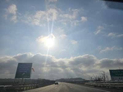The image displays a forward-facing perspective from a vehicle on a multi-lane asphalt highway under a partly cloudy sky. The sun is bright and centered in the upper frame, emitting significant lens flare. On the left side of the highway, a large green overhead sign reads "Çorlu", "Havaalanı" (Airport), "Çerkezköy", and "Tekirdağ" with an upward arrow indicating a straight direction and "400 m" below. A small red windsock-like object is affixed to the right edge of this sign. A partially visible green sign on the right side of the highway shows text including "Namık K..." and "Viyadük" (Viaduct), with an upward arrow and "429". Metal guardrails flank both sides of the roadway. Two light-colored passenger vehicles are visible in the far distance, traveling in the same direction on the highway. The background features distant low-lying hills with sparse vegetation. The scene depicts a highway environment in the vicinity of Ergene, Tekirdağ Province, Turkey, as indicated by the listed destinations on the road signs.