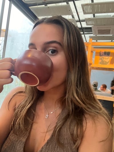 The image depicts a young woman consuming a beverage from a dark reddish-brown mug in an indoor establishment within Lordelo do Ouro e Massarelos, Portugal. She has long, light brown hair and wears a textured light brown top. A silver-colored necklace with a small pendant and a feather charm is visible on her chest. Her right eye is discernible, directed towards the camera, while the mug obscures her mouth and left eye. The background features a large, diffused-light window on the left, and on the right, wooden structures and a reflective surface display blurred figures of two male individuals, one wearing a cap and another with glasses. White rectangular ventilation grates and air conditioning units are mounted on the ceiling structure above.