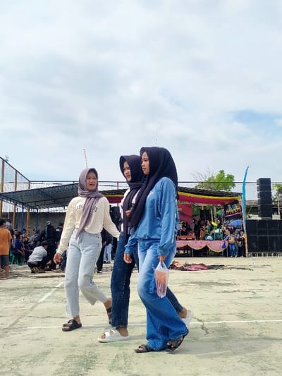 The image depicts an outdoor public gathering in Tamansari, Indonesia, under an overcast sky with scattered clouds. In the foreground, three young women, all wearing hijabs, walk from left to right across a light-colored paved surface marked with faded white court lines. The woman on the left wears a light purple hijab, a cream long-sleeved top with partial text, light blue wide-leg jeans, and black open-toed sandals; she is smiling and looking towards the camera. The central woman wears a black hijab, a white top, dark blue jeans, and white open-toed slides, looking towards her left. The woman on the right wears a black hijab, a light blue long-sleeved sweatshirt, blue wide-leg jeans, and dark open-toed sandals; she carries a clear plastic bag containing a reddish-brown beverage in her right hand and also looks towards her left. In the left background, a group of people, predominantly male, are gathered under a covered structure with a blue roof, secured by netting. In the right background, a stage adorned with pink and yellow fabric is visible, flanked by a tall stack of black audio speakers, with additional individuals congregated around and on the stage.