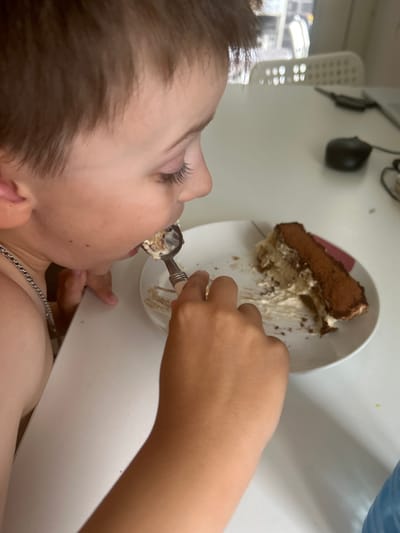 A close-up, overhead shot shows a young boy eating tiramisu from a white plate. He is using a spoon to take a bite of the dessert, which appears to be partially eaten. The boy has brown hair and is wearing a silver chain. In the background, a white wall is visible, along with some objects that are out of focus. The scene is likely indoors, possibly in a home setting. The lighting suggests it could be daytime.