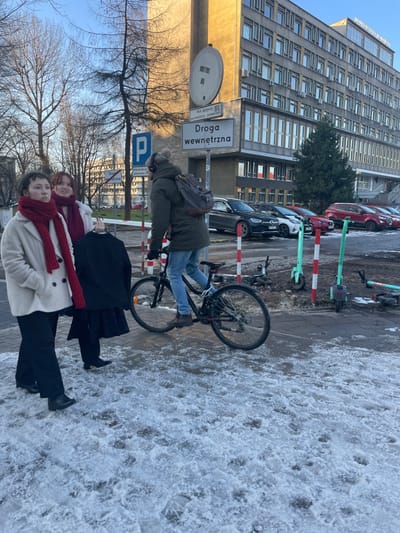 This photograph captures an urban winter scene in Kraków, Poland, under clear daytime skies. Three individuals are present on a snow-dusted sidewalk. In the middle ground, a person wearing a green jacket, blue jeans, and a grey hat with earflaps stands beside a black bicycle, their back partially turned, and a brown backpack visible. In the foreground, two women stand, both looking towards the camera. One wears a black coat and a red scarf, while the other is dressed in a light-colored coat with a matching red scarf, her reddish-brown hair visible.
The ground exhibits patches of dirty snow, ice, and slush, suggesting cold weather. Behind the people, several cars are parked along the street, and two green electric scooters are visible beside red and white striped poles. A prominent, multi-story building with a facade of numerous windows forms the backdrop on the left. Bare trees reinforce the winter setting.
Key urban elements include a large white rectangular street sign clearly reading "Droga wewnętrzna" (Polish for "Internal road"), a blue parking sign with a white 'P', and a partially obscured circular sign. The overall scene captures a crisp, mundane moment in a city during winter.