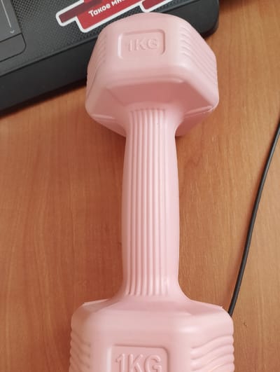 A pink dumbbell with "1KG" embossed on both ends rests on a wooden surface next to a black laptop and a black cord. The scene is indoors, likely a home or office environment. There is no information to suggest this scene is located in Birsk, Russia.