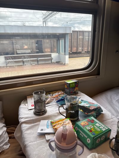 The image depicts an interior scene, likely inside a train carriage. A window on the left side shows an urban view, dominated by industrial structures and buildings. The scene appears to be shot in the city of Volkhov, Russia as there are inscriptions on the box in Russian and the "РЖД" mark on the train cup holder which means "Russian Railways".

On the table adjacent to the window are various objects, including two glass cup holders, the one on the lower left has inscription of "РЖД" which is the abbreviation for "Russian Railways", a box of "Tess" tea, snacks, and a children's educational game box. There is also a child's drinking cup with a lid.

The table is covered with a white, patterned tablecloth. Parts of the seating are visible in the background, suggesting the carriage is set up for passengers. The lighting is soft, indicating it's either an overcast day or a dimly lit environment.