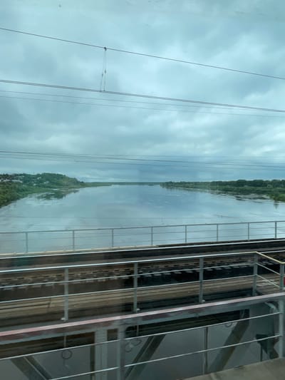 The image captures a view from a train, looking out over a wide river under an overcast sky. In the foreground, train tracks run horizontally across the frame, separated by a metal railing. The river occupies the midground and background, reflecting the cloudy sky and the lush green banks dotted with trees and some buildings on the left side. The scene appears to be in the city of Kotelnich, Russia, given the context provided. The overall atmosphere is calm and somewhat muted due to the cloudy weather.