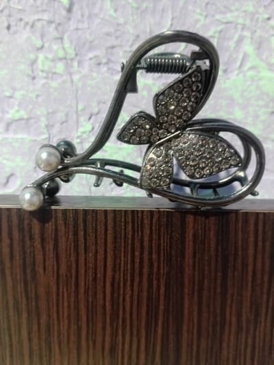 This media file features a close-up, eye-level shot of a decorative hair clip. The clip is shaped like a heart, with a butterfly design adorning its center. The butterfly's wings are encrusted with small, clear rhinestones, giving it a sparkling appearance. The heart shape itself is made of a dark, metallic material, possibly pewter or a dark-colored metal alloy. A coiled spring is visible within the heart's structure, suggesting the clip is functional. The clip is also embellished with two small, white pearl-like beads at the bottom.
The hair clip is positioned on the edge of a surface with a dark wood grain pattern, reminiscent of furniture or a door. The background is a textured, light-colored surface with a faint green tint, which appears to be a wall or fabric. The lighting suggests it is daytime, with the light source coming from the left, casting subtle shadows. The overall scene is simple and focuses on the intricate details of the hair accessory. There is no text visible in the image.