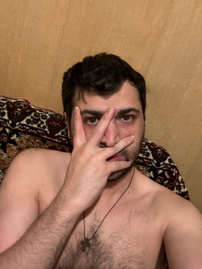A shirtless person with dark hair and a beard is lying down, partially obscuring their face with their hand. They are wearing a necklace with a cross pendant. The background consists of a wall with a wooden texture and a patterned fabric. The scene appears to be indoors, possibly a bedroom or living area, given the presence of a sofa or bed with a decorative blanket. The lighting is dim, suggesting it might be evening or night. There is no identifiable information indicating the location as Border Gyumri-Akhuryan, Armenia.