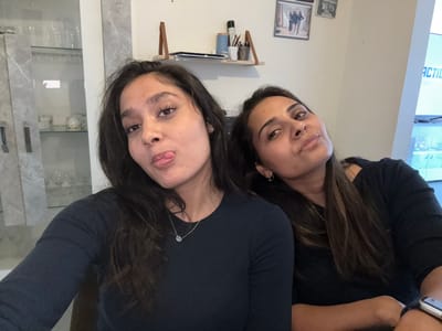 The image shows two women posing for a selfie indoors. The woman on the left has dark hair and is sticking out her tongue. She is wearing a dark long-sleeved shirt and a silver necklace. The woman on the right has long, dark hair and is looking at the camera with a slight smile. She is wearing a dark long-sleeved shirt and a silver necklace with a small pendant. They are seated, and the background includes a shelf with items like pens and cups, framed pictures on the wall, and a tall glass-fronted cabinet. There is also a television screen visible in the background. The scene appears to be a home environment. The location is identified as Maldegem, Belgium.