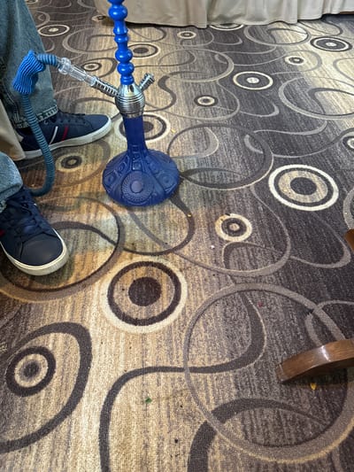 The image, taken from an overhead perspective, shows a person's legs and feet wearing dark blue tennis shoes with a white sole and red stripe. They are wearing blue jeans and are seated, with a blue hookah placed between their legs on a patterned carpet featuring concentric circles in tan, gray, and white. A partial leg of a wooden chair is visible in the upper right corner, and a section of white fabric is seen in the upper left corner. The location is Aygevan, Armenia, based on the prompt.
