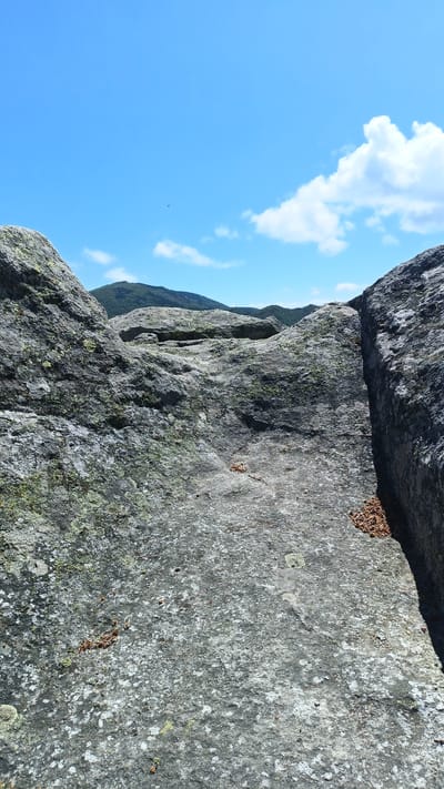 The image shows a rocky landscape in Sini Vrah, Bulgaria. Foreground features lichen-covered rocks with crevices. The background reveals rolling hills and a bright blue sky with scattered clouds. No people are visible. Some dried leaves are scattered across the foreground rocks.