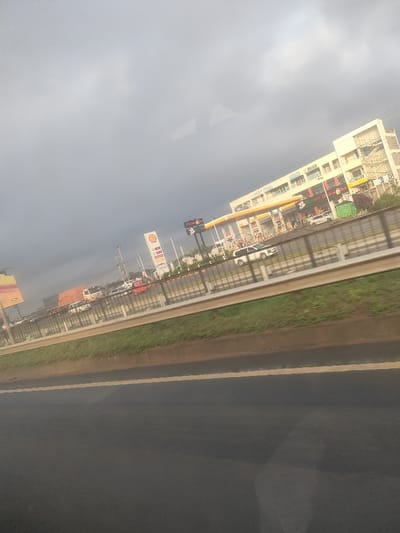 The image depicts a multi-lane highway scene in Murera ward, Kenya, under an overcast sky. In the foreground, a dark asphalt road surface with a faded yellow lane marking is visible, bordered by a brown curb and a strip of green grass. A metal guardrail runs horizontally across the mid-section of the frame, separating the viewer's immediate perspective from the opposing traffic lanes.

Beyond the guardrail, multiple vehicles are present, including a prominent white SUV, a red semi-trailer truck, and other cars, indicating moderate traffic flow. To the right, a Shell gas station is visible with its distinctive yellow canopy over fuel pumps and a tall red-and-yellow Shell logo sign. Directly behind the gas station stands a multi-story commercial building, predominantly white, with visible signage for businesses such as "Chicken Inn" and "Panda." Several utility poles are scattered throughout the midground. The background consists of a uniformly grey, overcast sky with indistinct distant structures. No specific individuals or discernible human actions are clearly identifiable.
