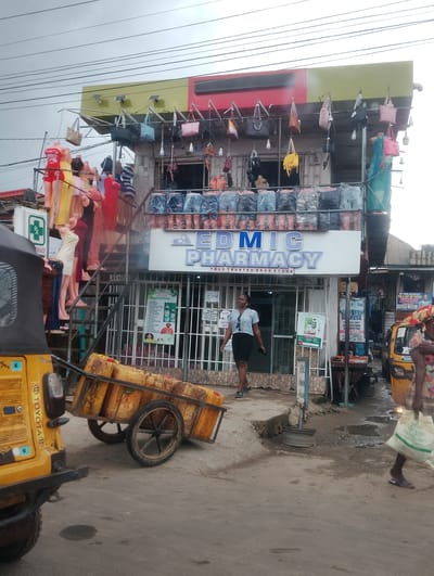 This is a full-shot, eye-level, outdoor photograph taken on a cloudy day in Iju, Nigeria. The scene depicts a street view of a business establishment with a pharmacy sign that reads "EDMIC PHARMACY YOUR TRUSTED DRUG STORE." The building has multiple levels, with various items displayed for sale. On the upper level, mannequins are dressed in clothing, and handbags are hung from the storefront. The lower level features storefront windows, and a woman in a striped shirt and dark skirt is walking past the entrance. To the left of the building, a yellow tricycle or auto-rickshaw is parked, and a cart filled with yellow jerrycans is being pushed. In the foreground, the street is paved with asphalt and shows signs of recent rainfall, with puddles of water visible. On the right side of the image, another woman is walking with a white bag. The overall environment suggests a bustling marketplace or commercial area.