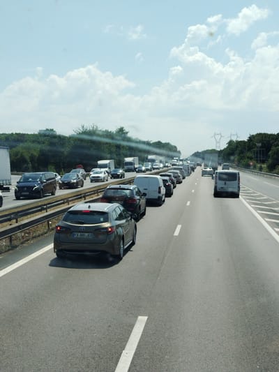 This image depicts a daytime scene of heavy traffic congestion on a multi-lane highway in Villeparisis, France. The sky is bright and partly cloudy, suggesting fair weather. The highway is lined with trees and vegetation on either side, and in the background, power lines and pylons are visible, indicating a semi-urban or rural setting.

The primary subjects are a large number of vehicles, including cars, vans, and trucks, all stalled or moving very slowly in a traffic jam. A dark grey station wagon with the license plate "FX-991-JC" is prominently featured in the foreground, facing away from the camera. Other vehicles in the traffic jam include a black Mercedes-Benz minivan, several sedans and SUVs, and multiple white panel vans and trucks. The vehicles are bumper-to-bumper, filling all visible lanes of the highway.

The activity taking place is a traffic jam, likely due to an accident, construction, or high volume of traffic on what appears to be a major road. There are no people clearly visible within the vehicles, but the sheer number of cars implies a significant number of occupants. The scene conveys a sense of frustration and delay typical of such traffic situations. The time of day is indicated by the bright sunlight and clear sky, likely mid-morning or afternoon. No specific emotional states are conveyed, as the focus is on the vehicles and the traffic situation.