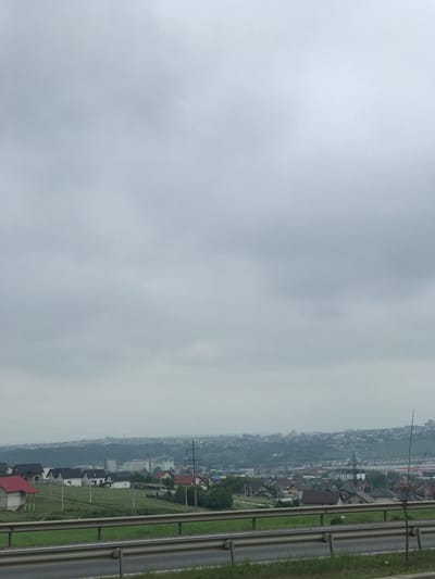 This image presents an expansive view of the town of Șcheia, Romania, under a uniformly overcast, light grey sky. In the immediate foreground, a metallic guardrail spans horizontally, with a narrow strip of asphalt road visible directly beneath it. A grassy embankment rises behind the guardrail, featuring several slender white posts and, on the right, a single thin, leafless sapling.

The midground reveals a scattered residential area comprising numerous single-family houses with diverse roof colors, including red, brown, black, and grey. Several houses exhibit photovoltaic solar panels on their roofs. Green vegetation, consisting of trees and bushes, is interspersed throughout the settlement, alongside visible utility poles and power lines. Further into the midground, a collection of larger, more densely clustered buildings, including a prominent light-colored industrial or commercial structure, indicates an area of mixed development.

The background consists of rolling hills blanketed with a combination of distant urban structures and dense foliage, appearing muted and somewhat indistinct due to atmospheric haze. No individuals or specific activities are discernible in the scene.